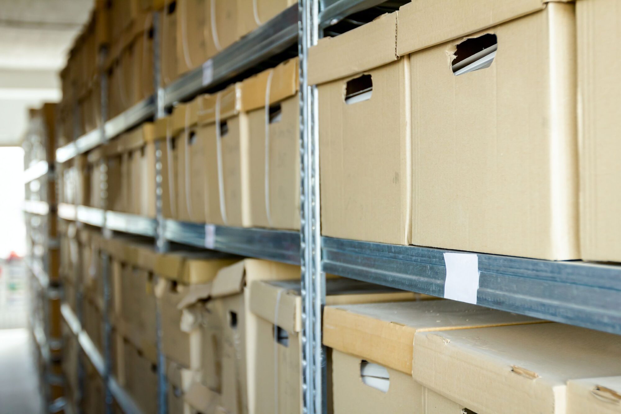 Cardboard archive boxes filled with documents are stored in shelving in a storage room.