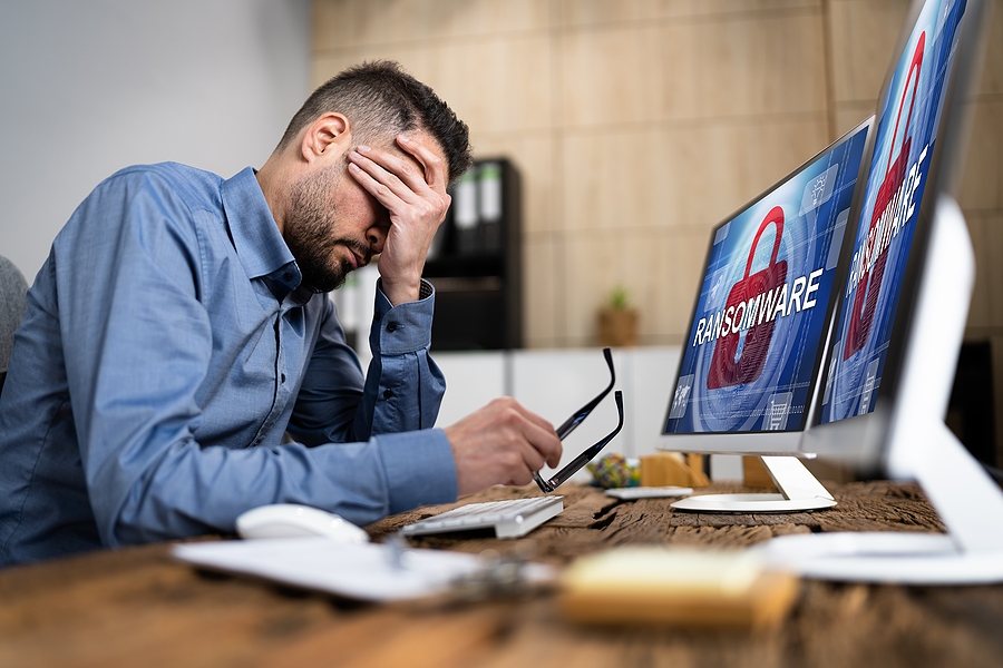 Stressed man holding his head at desk with computer screens showing a ransomware attack warning